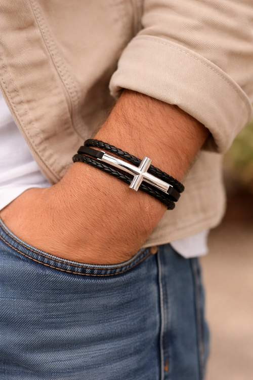 Close-up of a wrist wearing a black leather bracelet with a silver cross charm, with a blurred background.